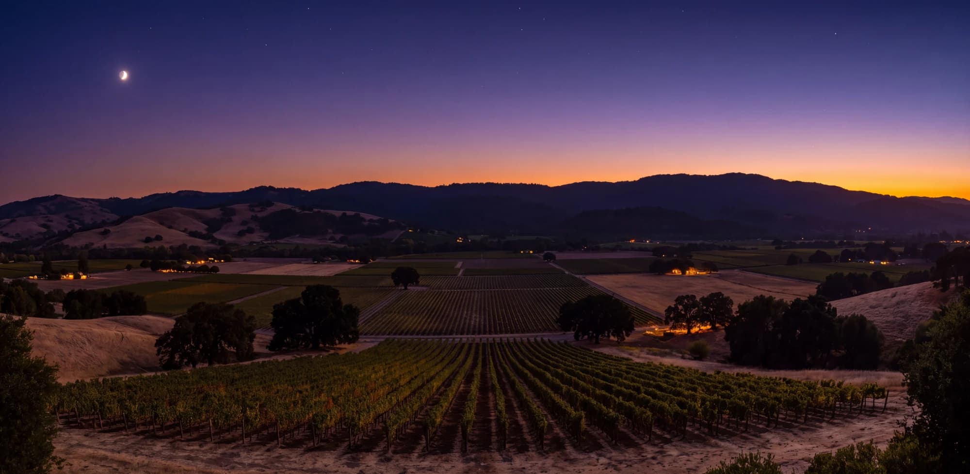 Dusk in the valley, a crescent moon over the ridge and the first lights on